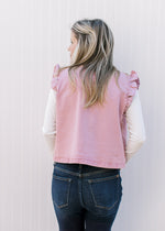 Back view of Model wearing a mauve denim vest with ruffle at arm and neck and slightly cropped fit.