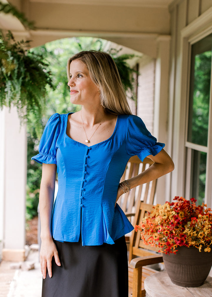 Model wearing a royal blue top with button-front, sweetheart neck, short sleeves and pleated hem.