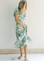 Model wearing a sage maxi dress with a cream floral design, pockets and a ruffle cap sleeve. 