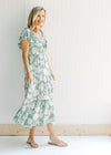 Model wearing sandals with a sage v-neck maxi dress with a cream floral design and a ruffle sleeve. 