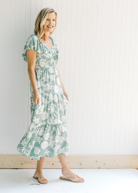 Model wearing sandals with a sage v-neck maxi dress with a cream floral design and a ruffle sleeve. 