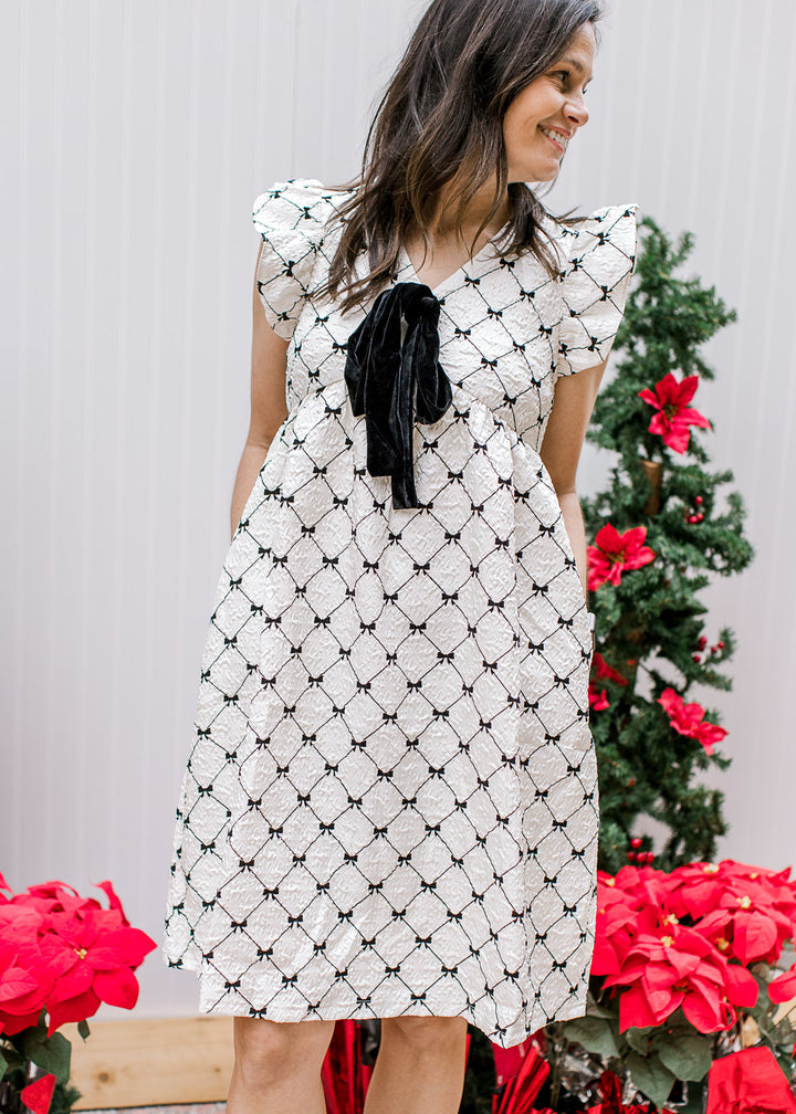 Model wearing a cream dress with black diamonds, v-neckline, pockets and flutter short sleeves. 