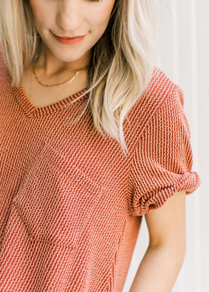 Close up of patch pocket and cuffed short sleeves on a v-neck rust colored tee. 