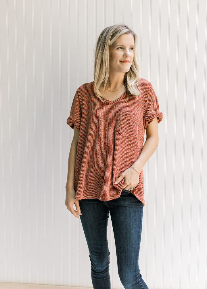 Model wearing jeans with a rust v-neck top with a corded material, patch pocket and cuffed sleeves. 