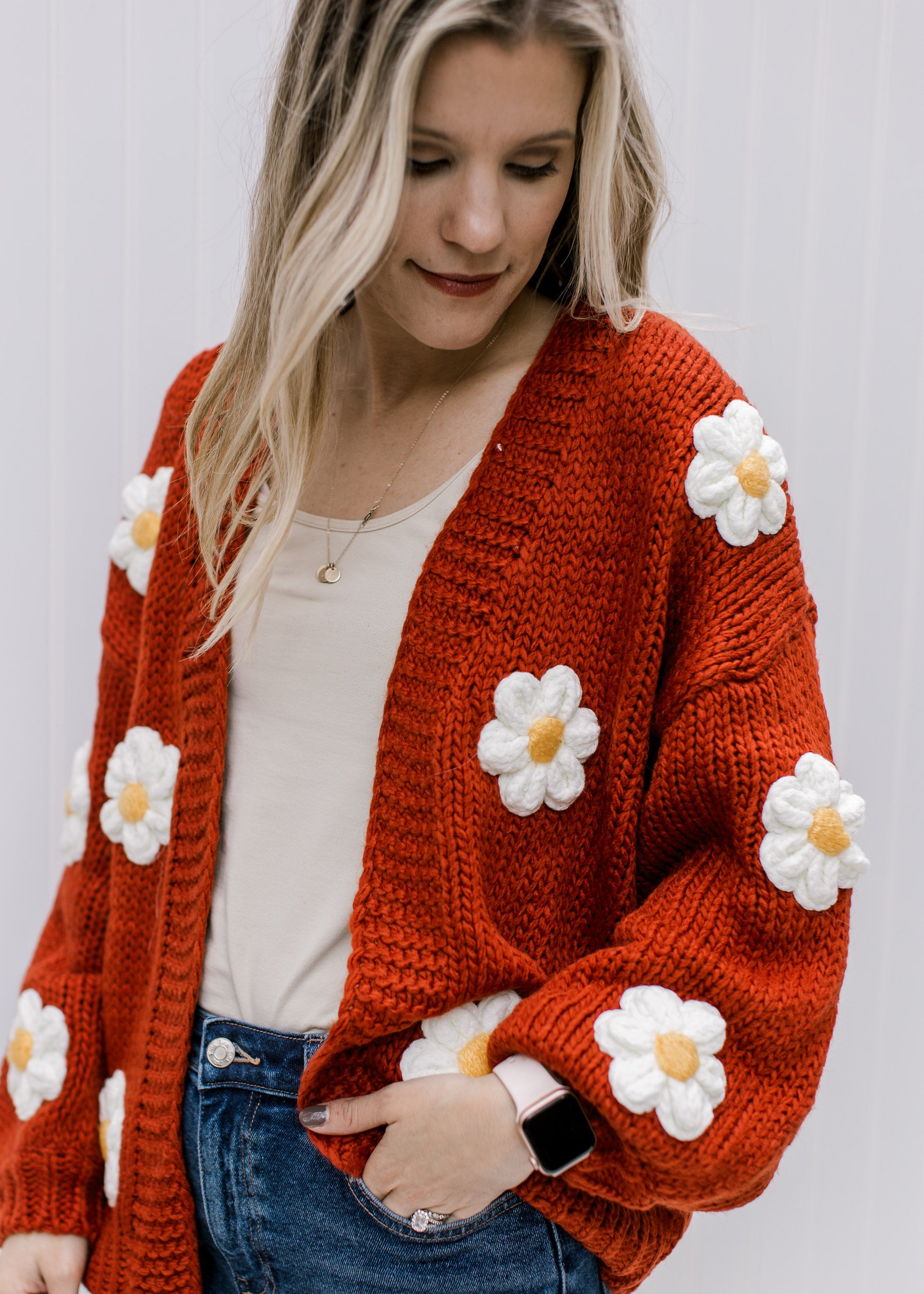 Model wearing a rust cardigan with white and yellow embroidered daisies and long sleeves. 