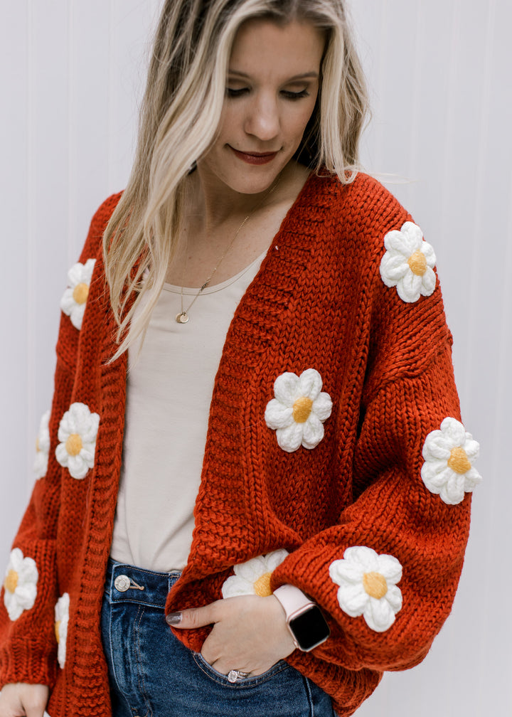 Model wearing a rust cardigan with white and yellow embroidered daisies and long sleeves. 