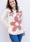 Model wearing a cream top with rust colored stitched flowers, long sleeves and a round neck. 