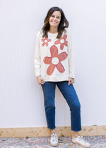 Model wearing jeans with a cream top with rust stitched flowers, long sleeves and a round neck. 