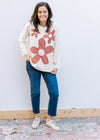 Model wearing jeans, sneakers and a cream long sleeve top with rust colored stitched flowers. 