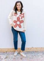 Model wearing jeans, sneakers and a cream long sleeve top with rust colored stitched flowers. 