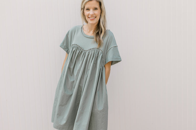 Model wearing a sage above the knee dress with exposed hem, short sleeves and a ruffle detail.