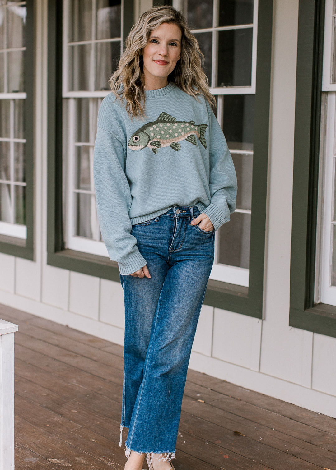 Model wearing flats, jeans and a light blue sweater with a sage fish on front and long sleeves. 