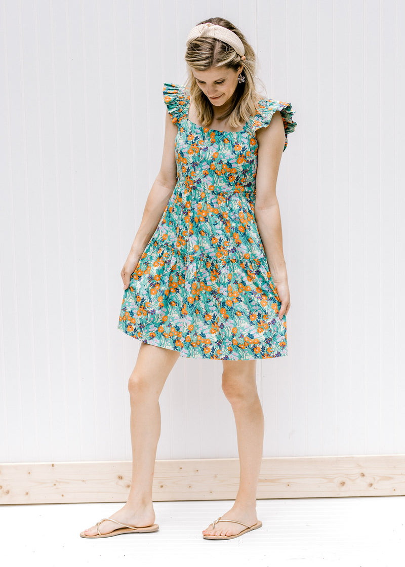 Model wearing a tiered sage floral dress with a square back and ruffle sleeves and sandals. 