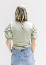 Back view of a Model wearing a sage top with a ruffle detail on a short sleeve and a round neck. 