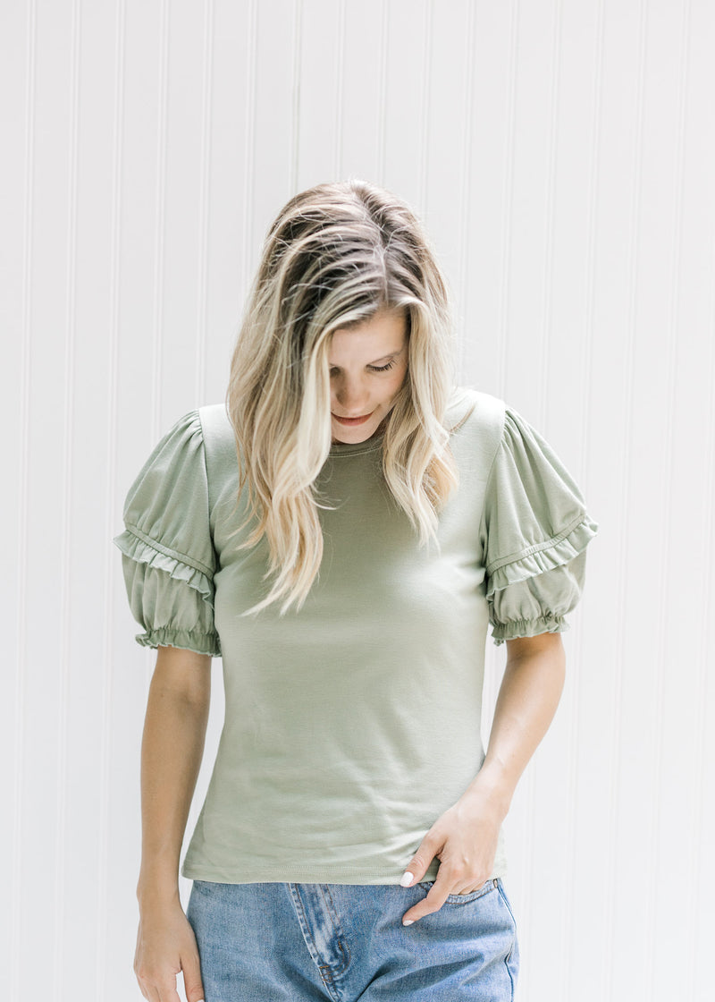 Model wearing jeans with a sage top with a ruffle detail on a short puff sleeve and a round neck. 