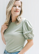 Model wearing a sage top with a ruffle detail on a short puff sleeve and a round neck. 