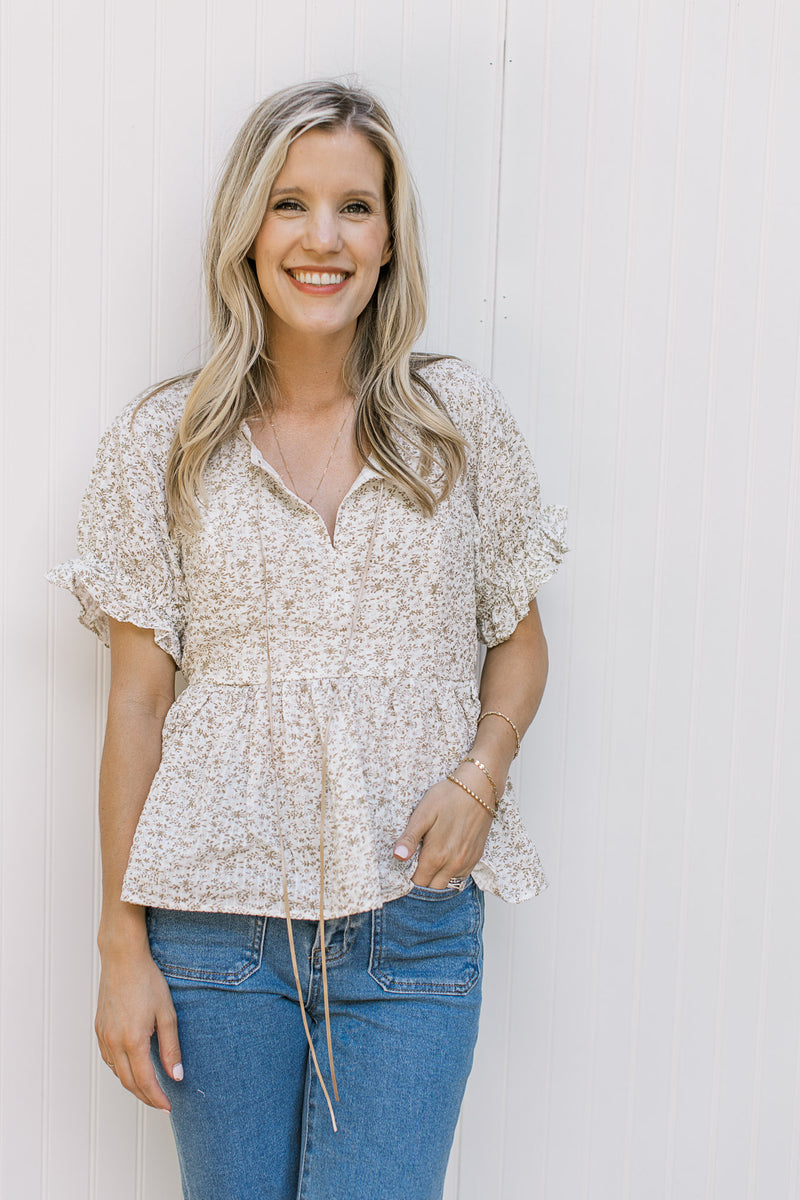 Model wearing jeans with a white top with sand flowers a v-neck with tie detail and a peplum cut.