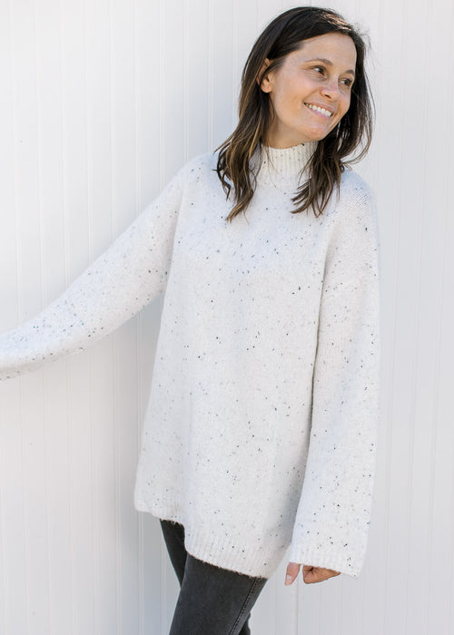 Model wearing an oversized off white sweater with black flecks, long sleeves and a mock neck. 