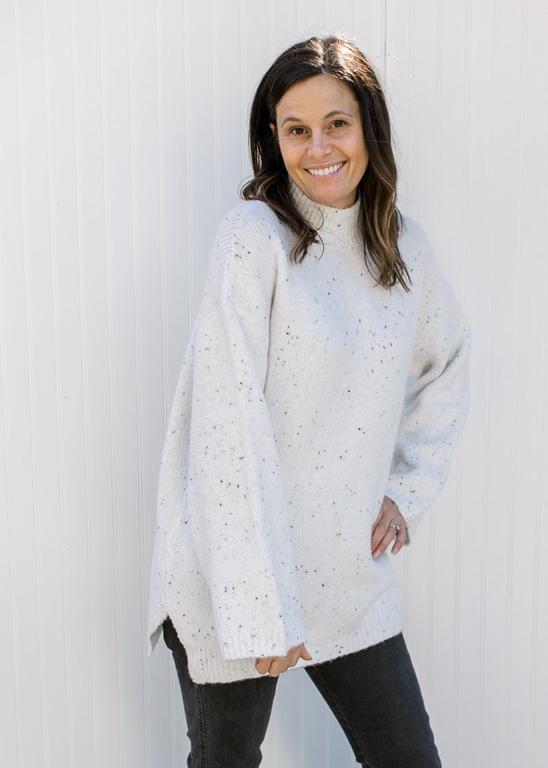 Model wearing an oversized off white sweater with black flecks, long sleeves and split sides.