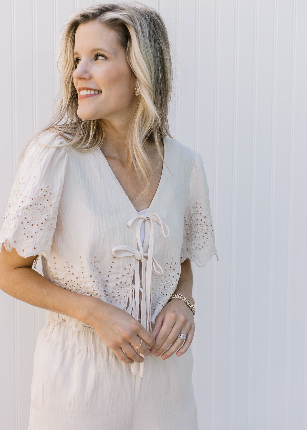 Close up of a cream, short sleeve, v-neck  top with scalloping and eyelets and three tie closure.