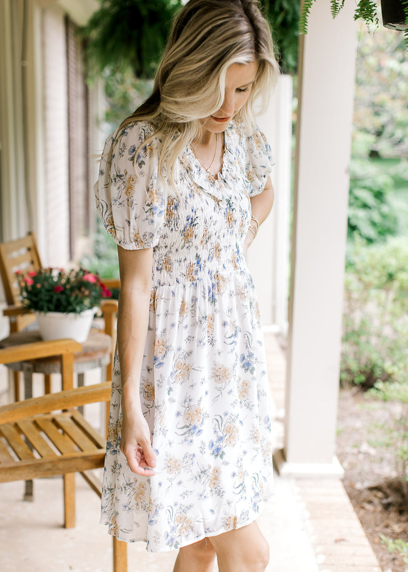 Model wearing a white above the knee dress with yellow and blue floral and a smocked bodice. 