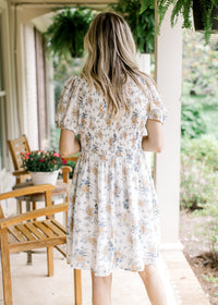 Back view of Model wearing a white dress with floral, v-neck, bubble sleeves and a smocked bodice. 
