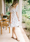 Model wearing sandals and a white V-neck dress with yellow and blue floral and a smocked bodice. 