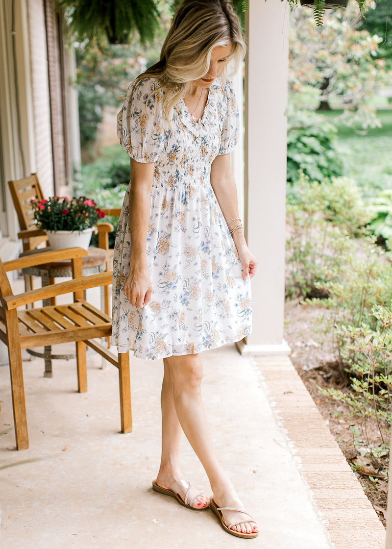 Model wearing sandals and a white V-neck dress with yellow and blue floral and a smocked bodice. 