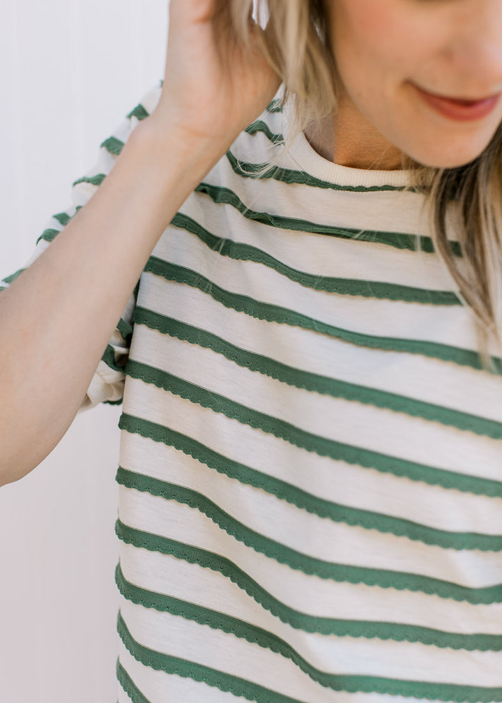 Model wearing a cream top with green scalloped stripes, short sleeves and a round neck.
