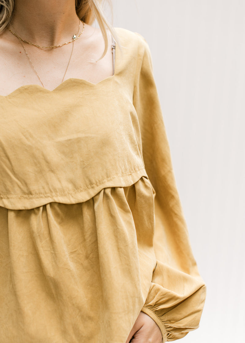 Close up of scalloped detail at neck and breast on a mustard colored top with long sleeves. 