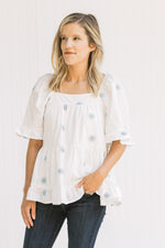 Model wearing a white top with blue embroidered daisies, a square neckline and bubble short sleeves.