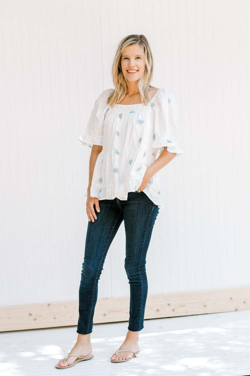 Model wearing jeans with a white top with blue embroidered daisies, a square neck and short sleeves.