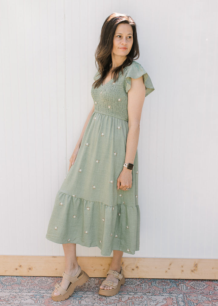 Model wearing wedge sandals and a sage green midi with cream embroidered flowers. 