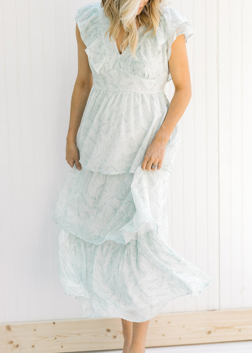 Model wearing a white, tiered, v-neck maxi dress with a green leaf pattern and ruffle cap sleeves. 
