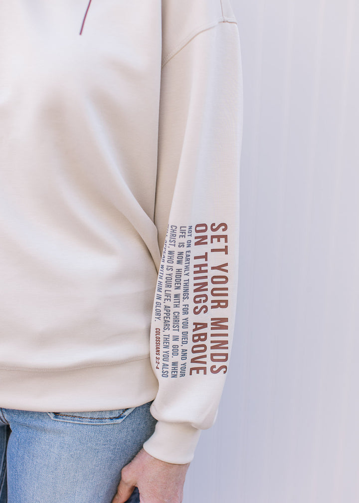 Close up of “Colossians 3:2–4” on left long sleeve of a cream pullover with a round neckline. 