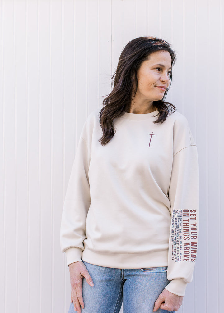 Model wearing a cream pullover with maroon cross detail and “Colossians 3:2–4” on left sleeve. 