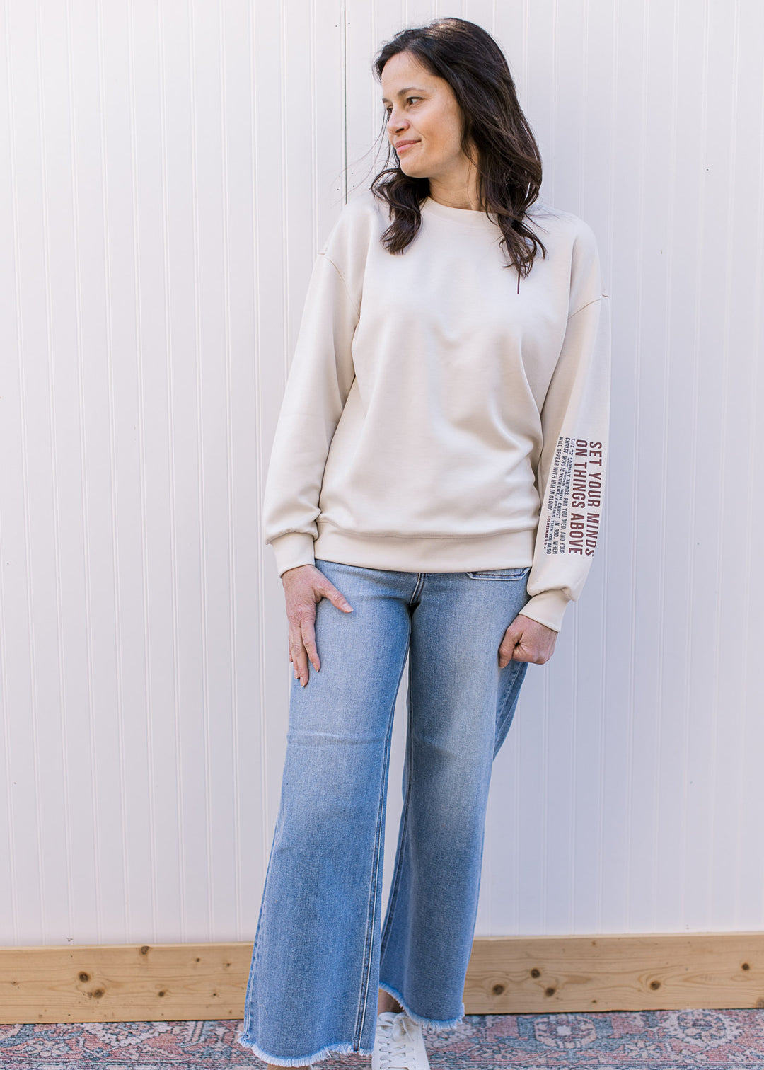 Model wearing jeans and a cream pullover with maroon cross and “Colossians 3:2–4” on left sleeve. 
