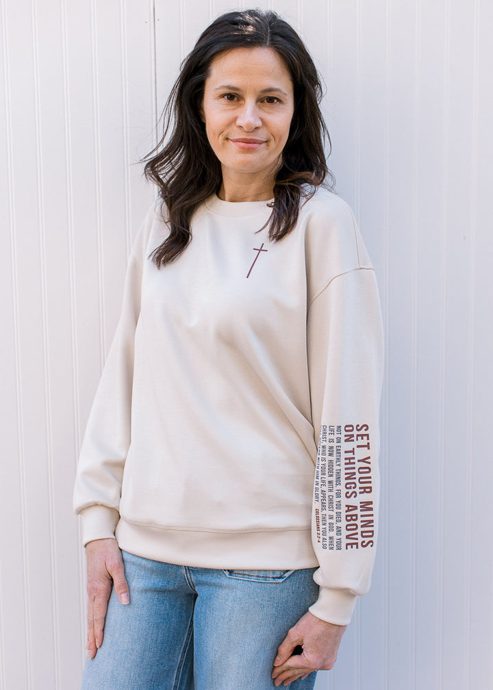 Model wearing a cream top with maroon cross detail round neck and “Colossians 3:2–4” on sleeve. 
