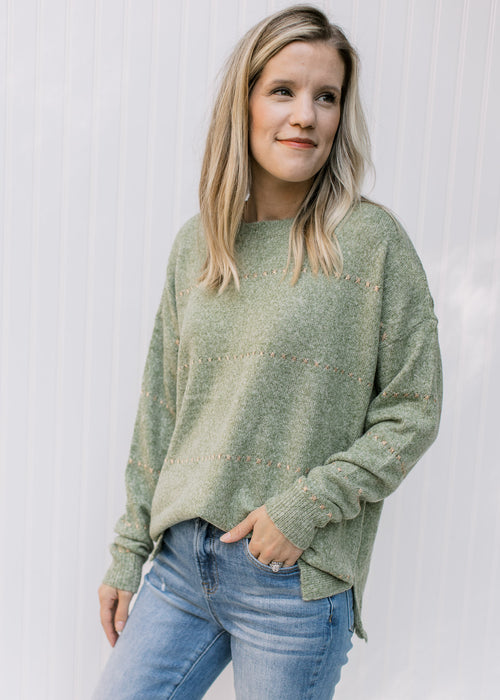 Model wearing a mottled green knit sweater with gold accent stitches and a ribbed neck and sleeve. 