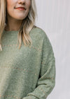 Close up of mottling on a green knit sweater with a round neck and gold accent stitching. 