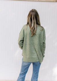 Back view of Model wearing a mottled green knit sweater with gold accent stitches and long sleeves. 