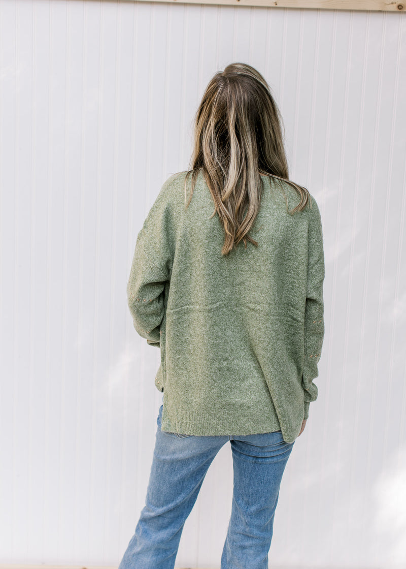 Back view of Model wearing a mottled green knit sweater with gold accent stitches and long sleeves. 