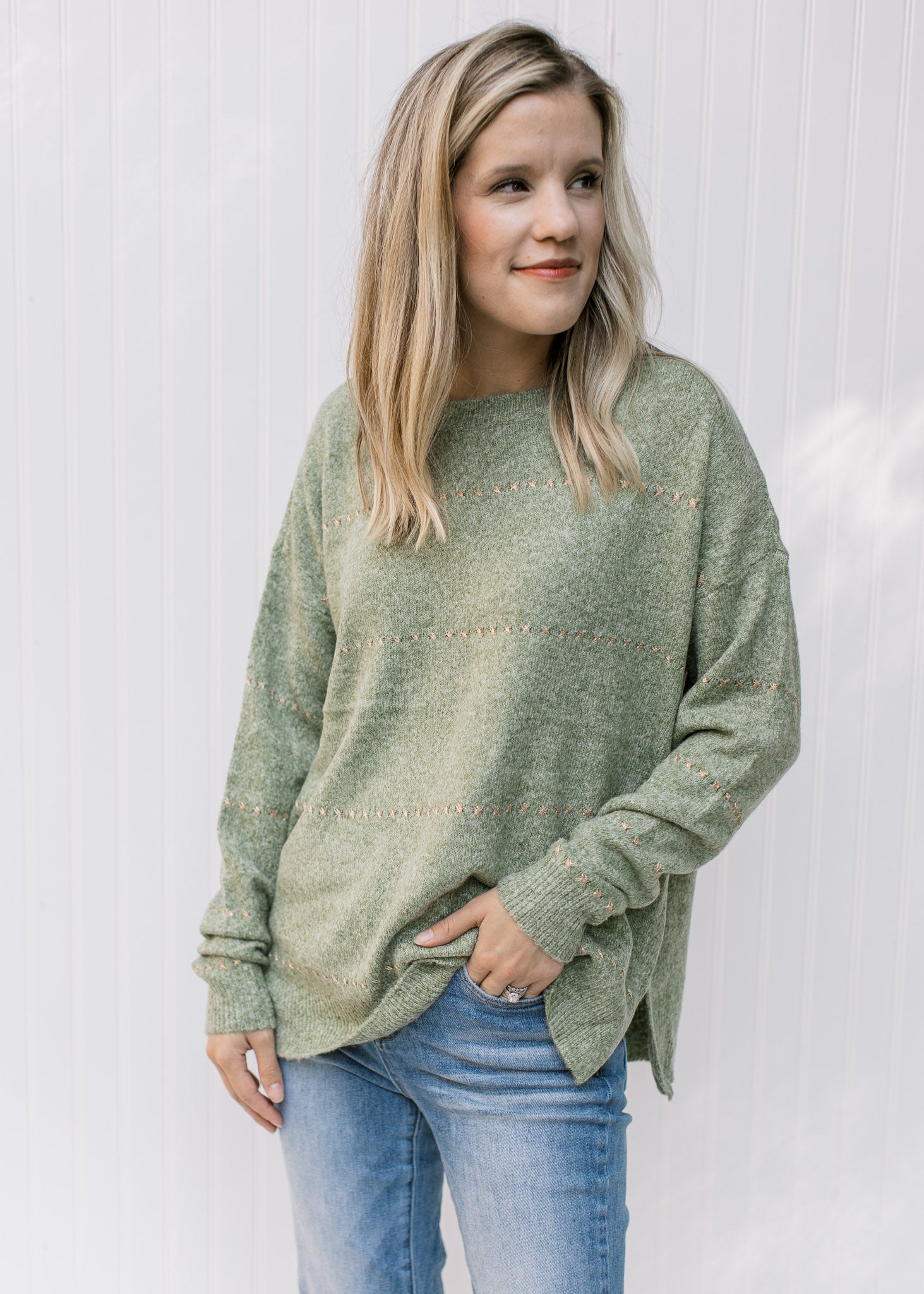 Model wearing jeans with a mottled green knit sweater with gold accent stitches and long sleeves. 