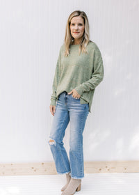 Model wearing jeans and booties with a mottled green sweater with gold stitching and long sleeves. 
