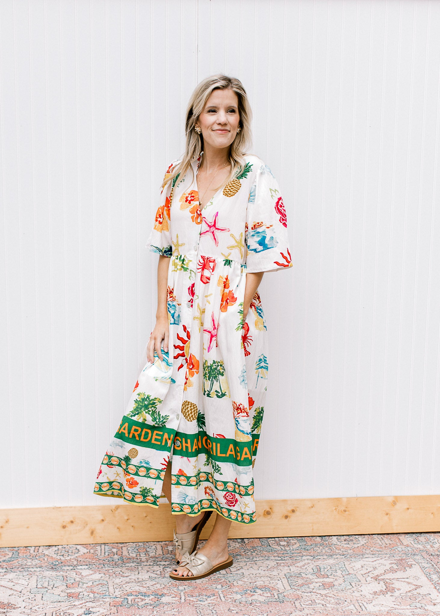 Model wearing sandals and a button up cream midi with a v-neck and multicolored tropical pattern. 