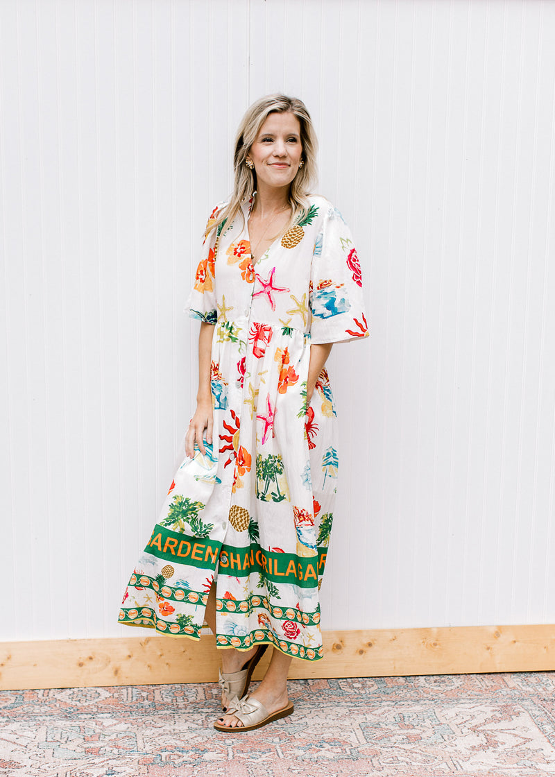 Model wearing sandals and a button up cream midi with a v-neck and multicolored tropical pattern. 