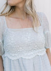 Close up of lace overlay at the bodice of a pale blue short sleeve top with a square neckline. 