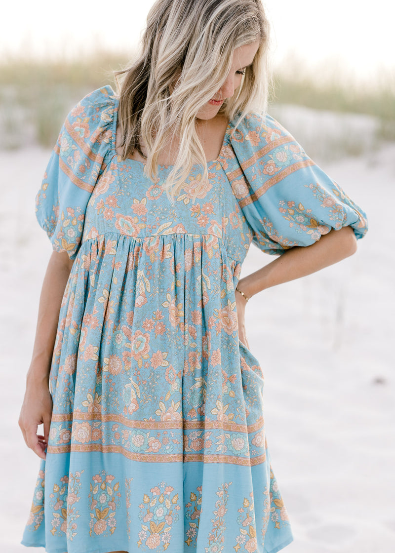 Model wearing a light blue above the knee dress with a gold floral pattern and short sleeves.