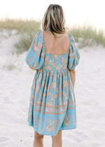 Back view of Model wearing a light blue dress with a gold floral pattern and square neck. 