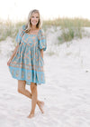 Model wearing a light blue, above the knee, dress with a gold floral, square neck and short sleeves.